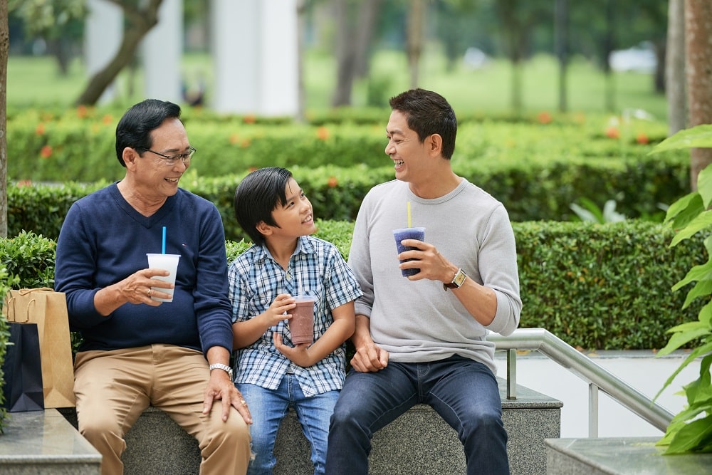 Asian boy smiling and enjoying tasty beverage while sitting between father and grandfather in park